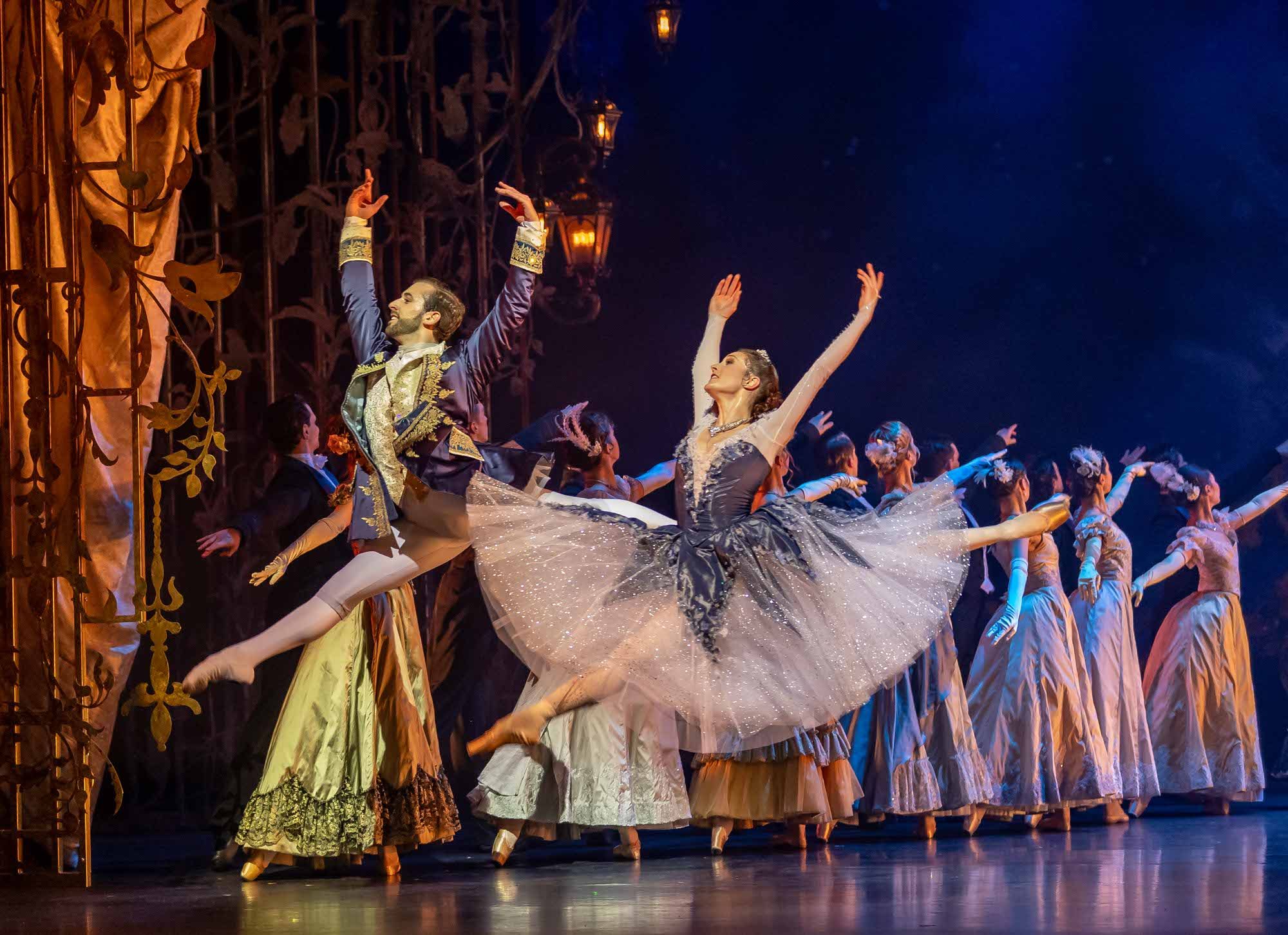A male and female ballet dancer leap gracefully on stage in elegant costumes, surrounded by other dancers in period attire and warm lighting, creating a dramatic and enchanting performance scene.